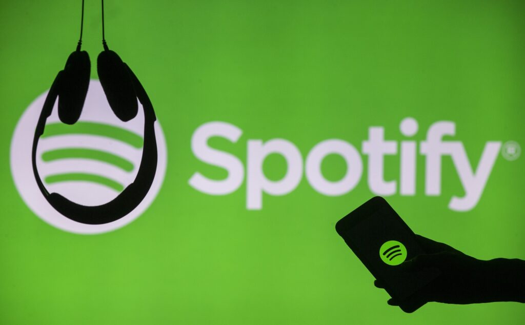 logo di spotify con davanti in controluce l'immagined i una mano che regge uno smartphone e due cuffie appese